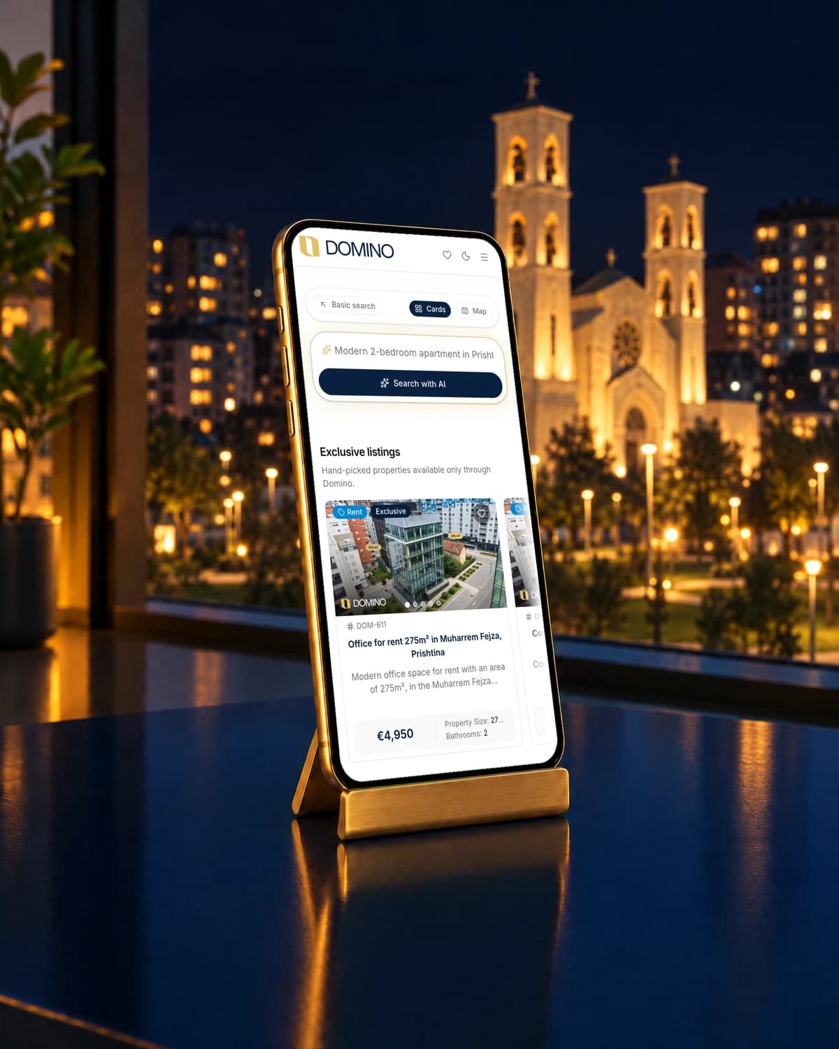 A phone on a brass stand showing the Domino website, with a floodlit cathedral in Prishtina behind