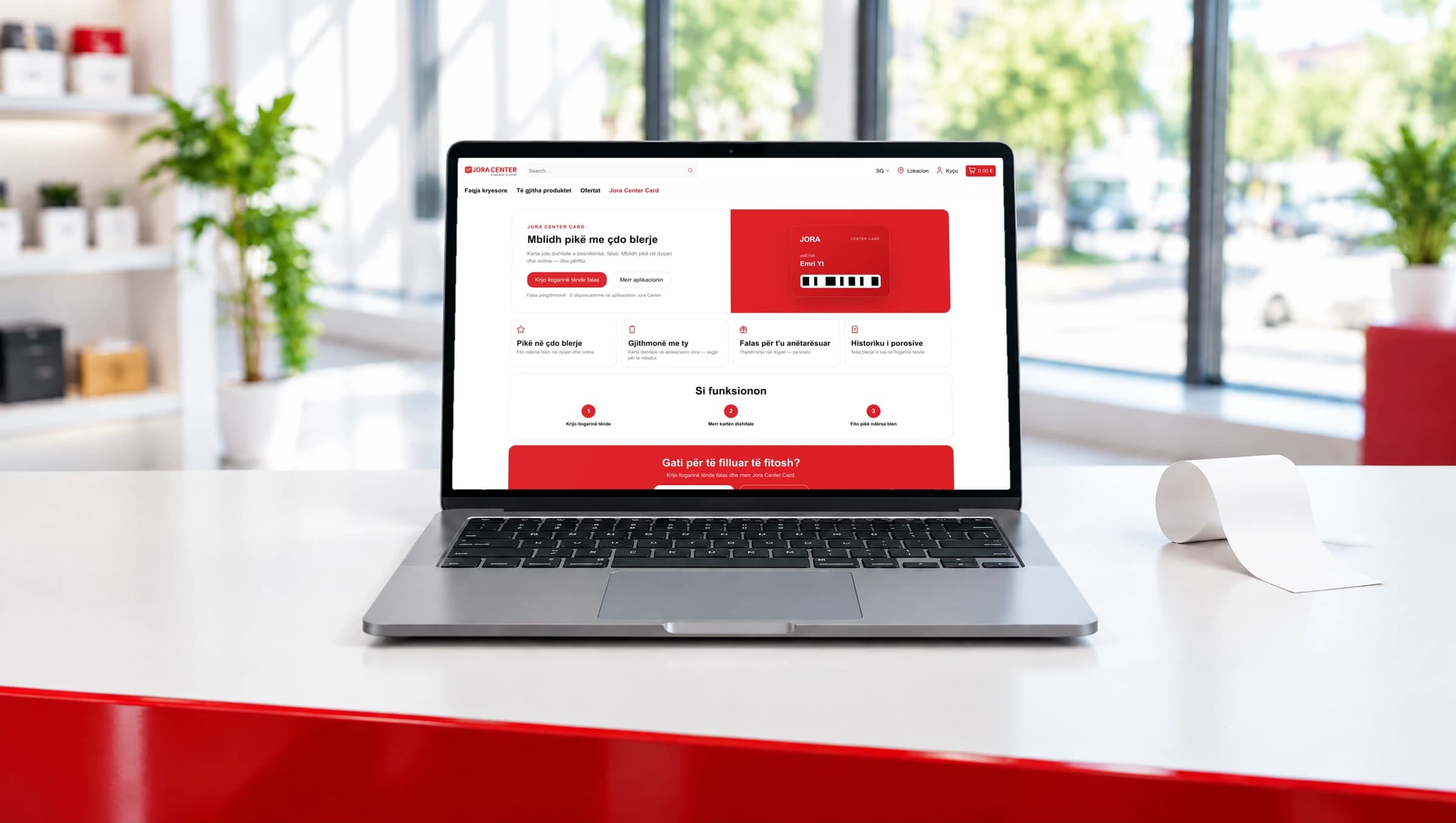 Ein Laptop auf einer langen weissen Ladentheke mit roter Kante zeigt die Seite zur Jora Center Card, daneben ein gekräuselter Streifen leeres Quittungspapier