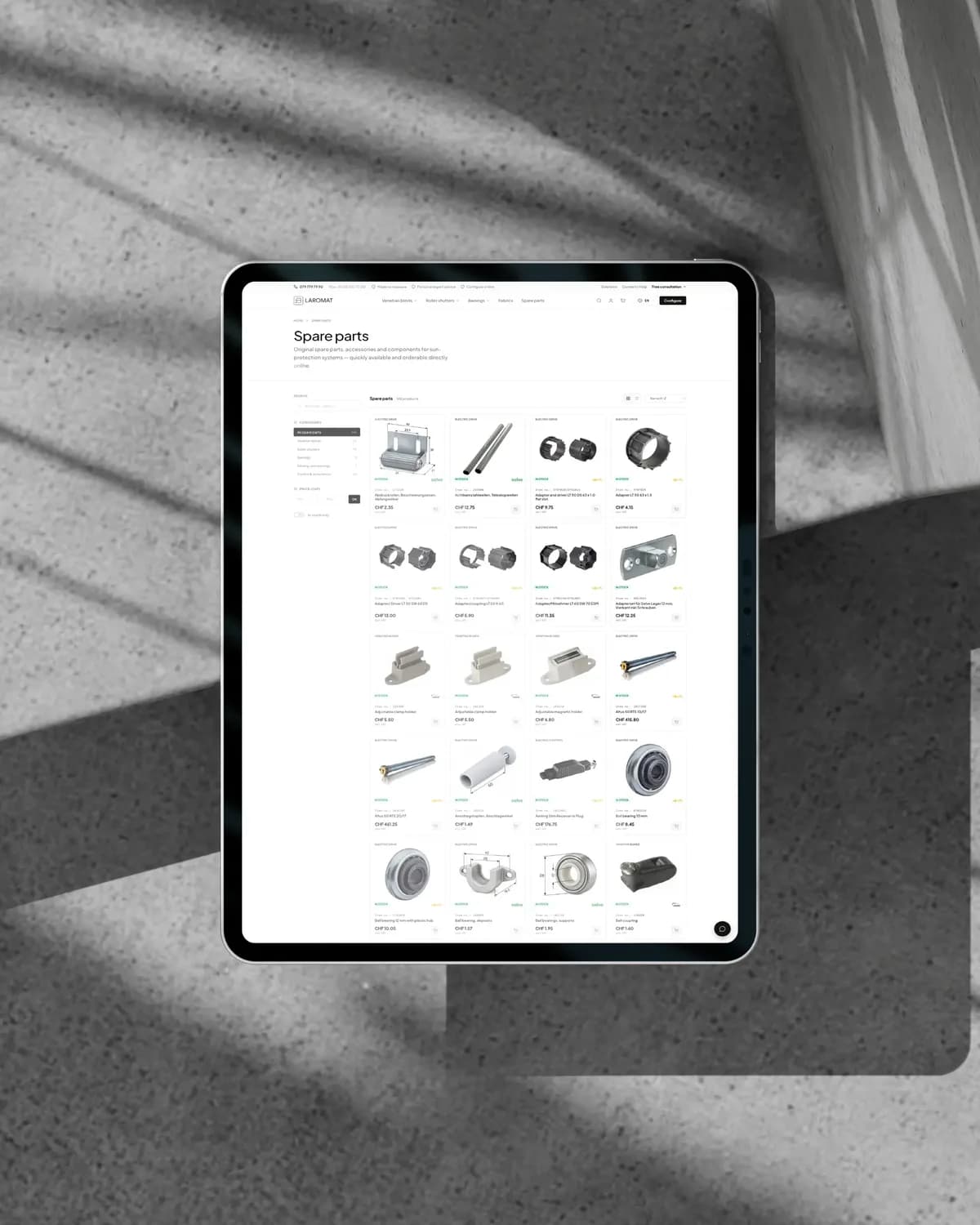 Ein Tablet auf Beton im Licht durch Lamellen, darauf der Ersatzteilkatalog von Laromat mit Kategoriefilter und einem Raster von Teilen mit Preisen
