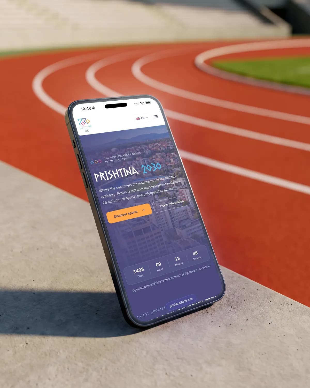 A phone leaning on a concrete kerb beside a stadium running track, showing the Prishtina 2030 home page with the countdown to the Games