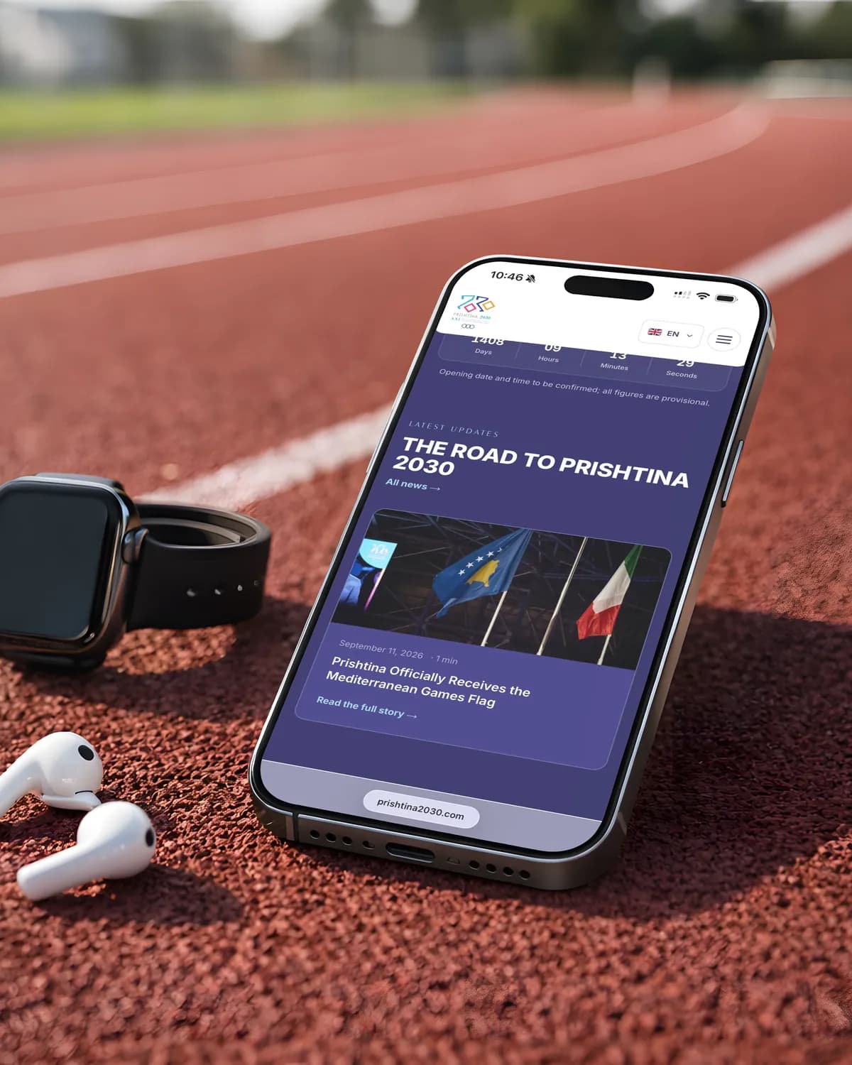 A phone on a running track beside a smartwatch and earbuds, showing the Prishtina 2030 news: The road to Prishtina 2030, with the story on receiving the Mediterranean Games flag