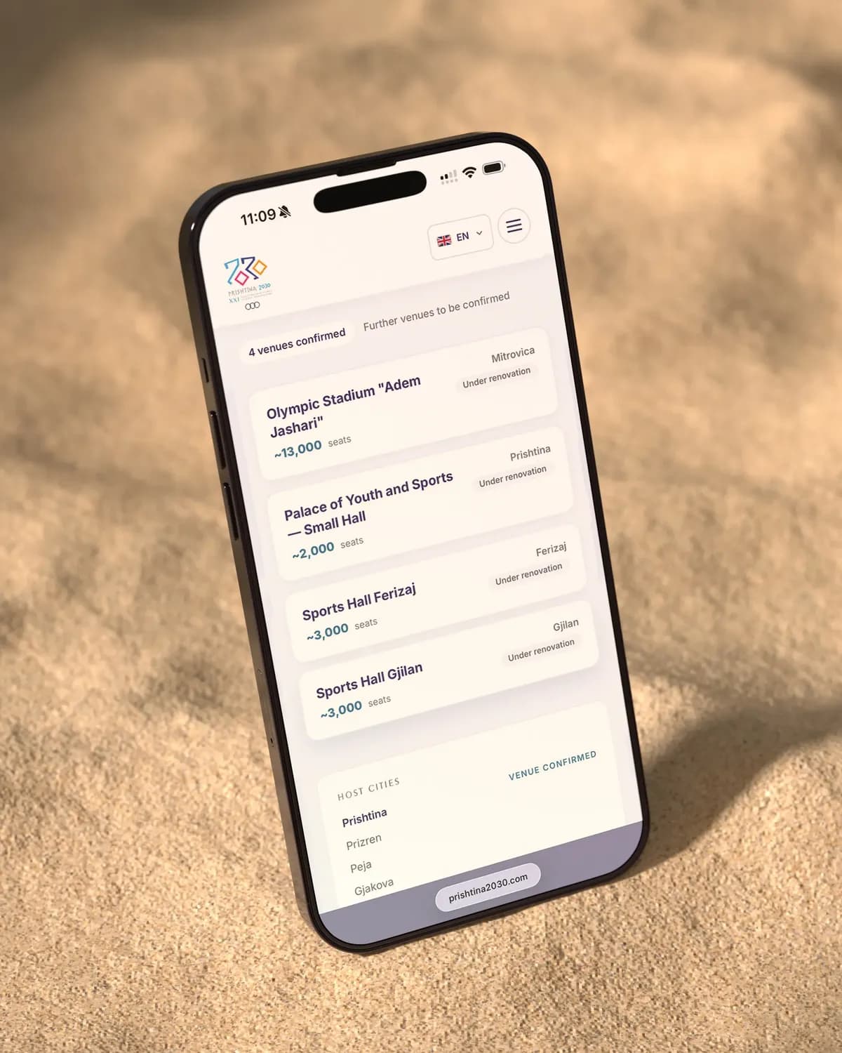 A phone standing in sand, showing the Prishtina 2030 venues: the venues confirmed so far with their seats, and the host cities